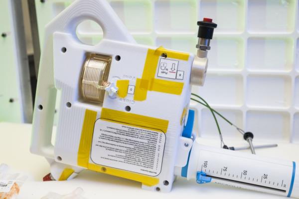 The image shows a close-up of the Mini Potable Water Dispenser prototype. The device is primarily white with yellow tape on it and has several instructional labels on its surface. It features a prominent graduated syringe-like component for precise water measurement, marked in milliliters, and a black push button at the top. The device has a handle on one side for easy portability. The background consists of a white grid pattern, which helps draw attention to the device. Partially visible in the foreground are vacuum-sealed food pouches, indicating the device's use for rehydrating food.