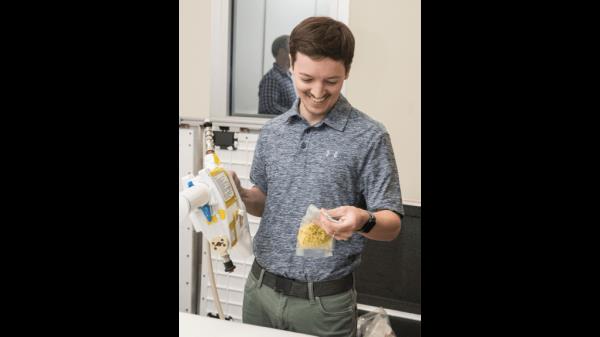 The image shows a close-up of a person filming a demonstration with a smartphone. The scene being recorded involves the use of a Mini Potable Water Dispenser to rehydrate a vacuum-sealed food pouch. The person's hand is holding the device while directing water into the pouch, which is positioned on a table. The smartphone's screen is clearly visible in the foreground, displaying a live video of the demonstration in progress